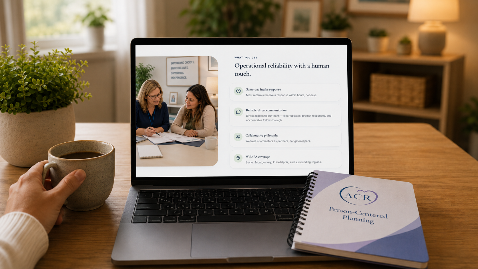 A laptop on a warm wooden desk showing the Absolute Care Residences site, next to a coffee mug and an ACR Person-Centered Planning notebook